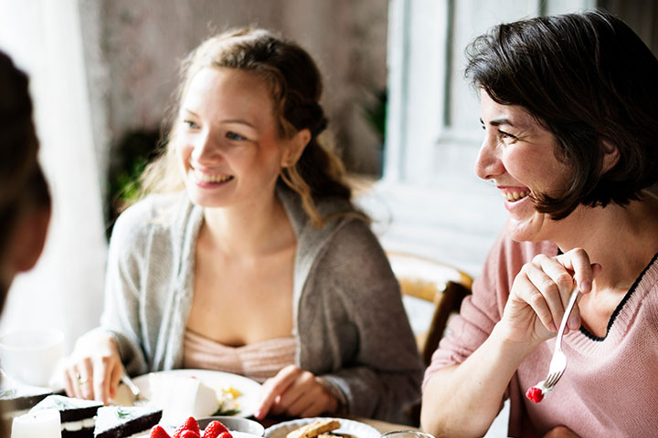 Zwei Frauen lachen und genießen ein Essen in einem hellen Raum. Eine hält eine Gabel mit Essen.