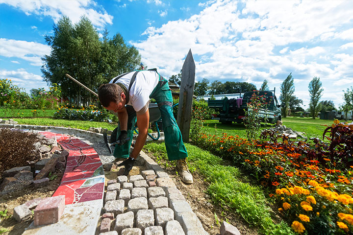 Arbeiter in grüner Kleidung verlegt Pflastersteine auf einem Gehweg neben einem farbenfrohen Blumenbeet.